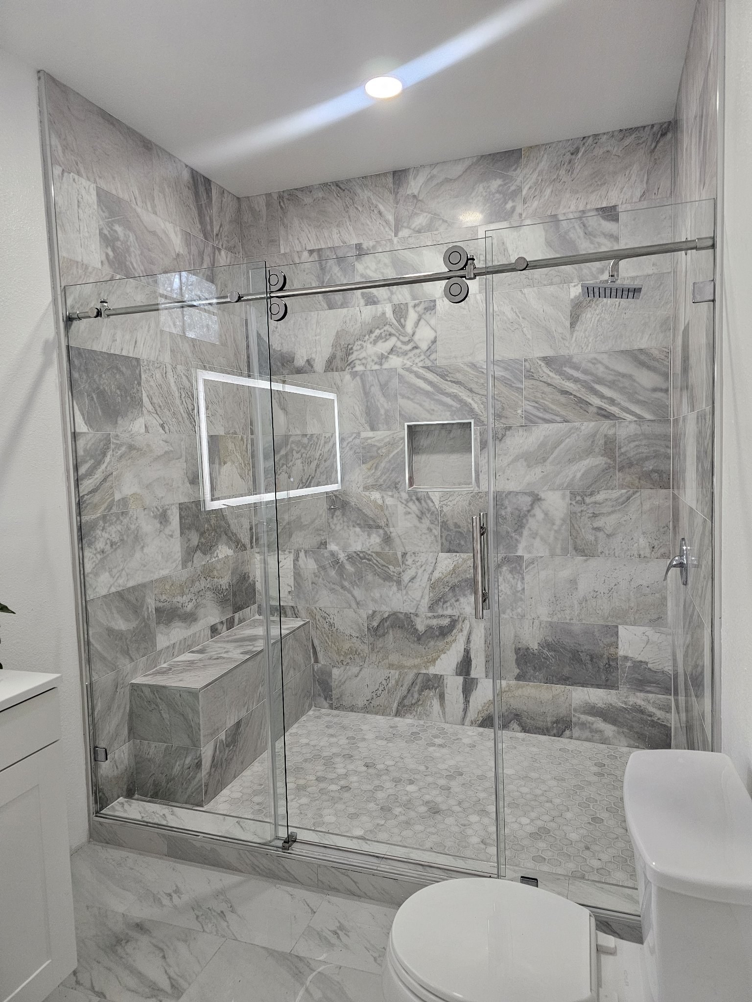 Sliding de una puerta con panel fijo y riel de brush nickel en shower de mármol gris con banca y nicho